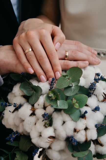 Close-up of a newlywed couple's hands, both wearing gold wedding bands, gently resting on a floral arrangement. The bouquet features soft, fluffy white cotton bolls, accented with sprigs of aromatic b