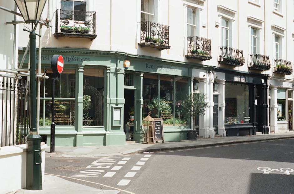 A light green floral boutique situated on a corner of a street with large display windows showcasing fresh flowers and potted plants. The shop has elegant black wrought-iron balconies above, decorated
