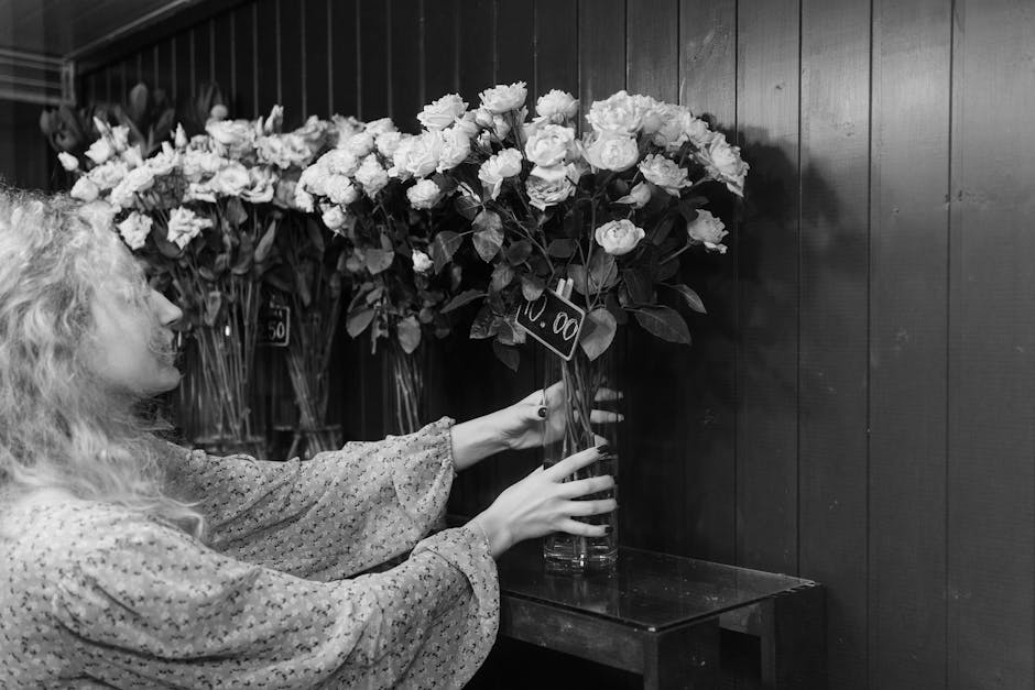 A woman with curly blonde hair, wearing a patterned long-sleeve blouse, is arranging a large bouquet of fresh white roses and greenery in a tall glass vase. She is holding the vase with both hands, po